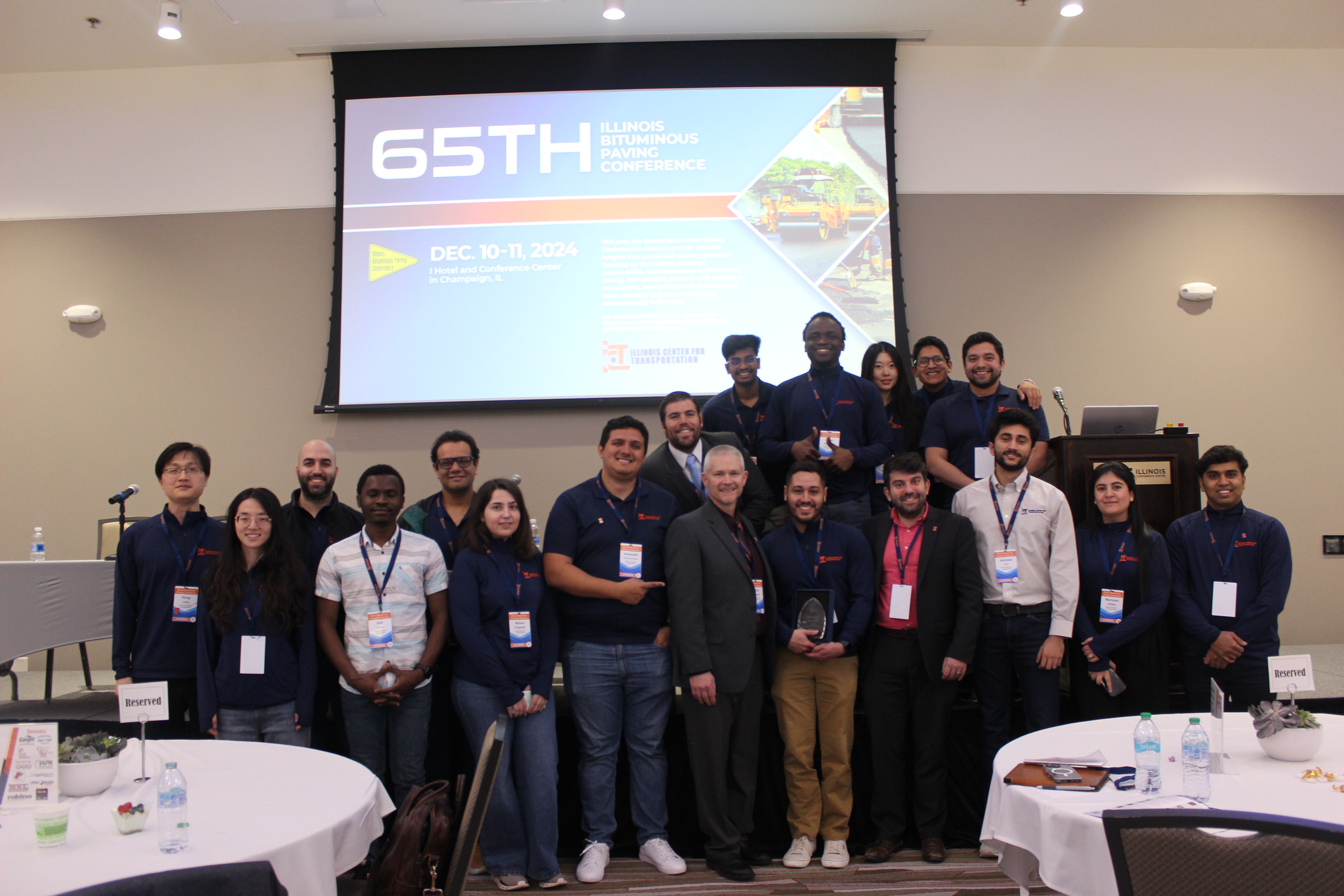 A group of ICT staff and students pose in front of a display that says 65th Illinois Bituminous Paving Conference.